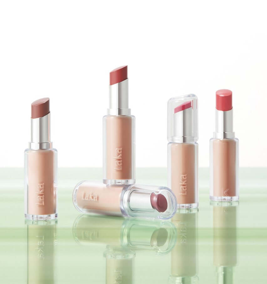 Laka Bonding Glow Lipstick collection showcasing luxurious colors in clear tubes on a smooth green surface.