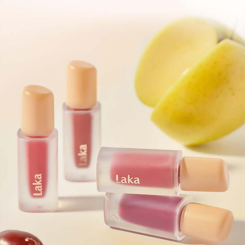 Laka Fruity Glam Tint lip tints in clear tubes surrounded by fresh fruit, showcasing juicy color and high-shine gloss.