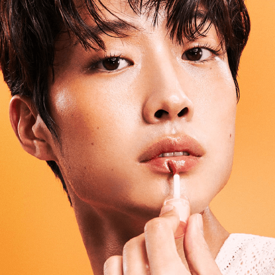 Person applying Laka Jelling Nude Gloss for glossy, hydrated lips in a vibrant orange background.